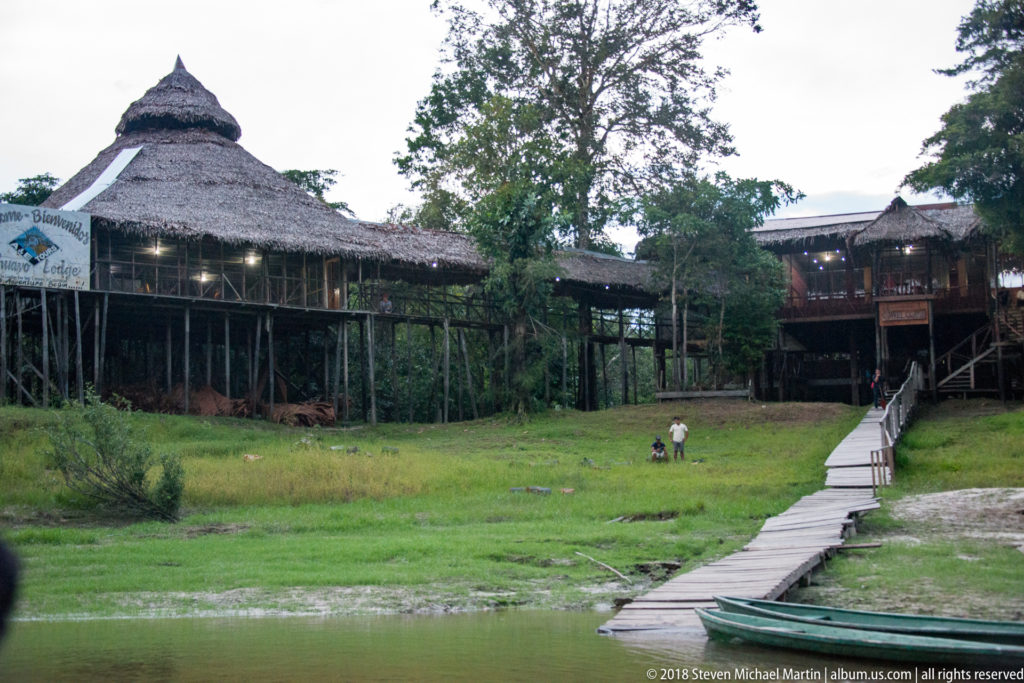 Long boardwalk leading from the riverbank to large palm-thatched lodge buildings raised on stilts around a grassy clearing.