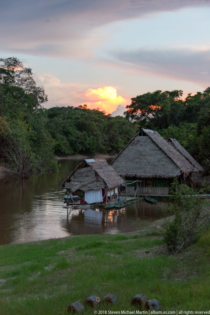 Large palm-thatched riverside building with smaller shelters and moored boats beneath an orange sunset horizon.