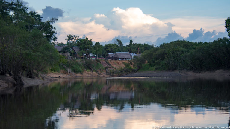 Forest-lined river bend reflecting large clouds, with a small settlement visible on the distant bank.