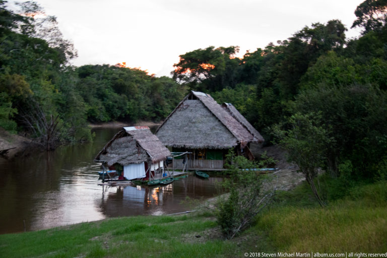 Close view of palm-thatched riverside buildings with moored narrow boats on a calm Amazonian channel at dusk.