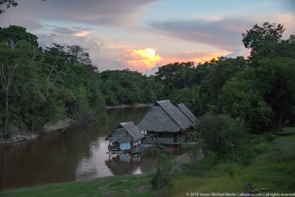 Palm-thatched riverside buildings and small boats below a glowing sunset, surrounded by dense Amazonian forest.