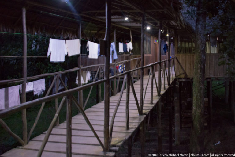 Elevated palm-thatched wooden walkway with rustic railings, hanging white laundry, and lights glowing along lodge rooms.