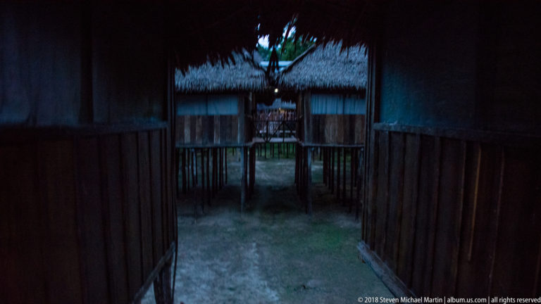 Dark wooden passage at night framing a softly lit palm-thatched lodge building and veranda beyond.
