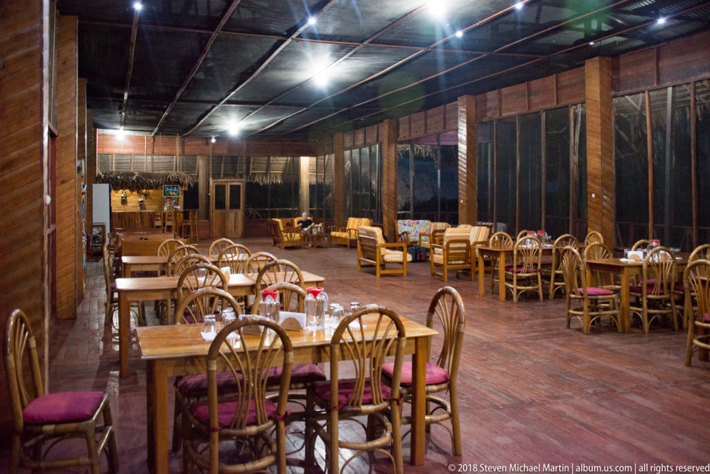 Large wooden dining room with many tables, curved chairs, screened walls, and bright ceiling lights at Tuhauyo Lodge.