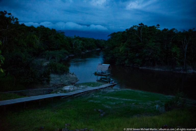 Dark river channel at dusk beside dense forest, with a roofed landing, moored boats, and long walkway in the foreground.