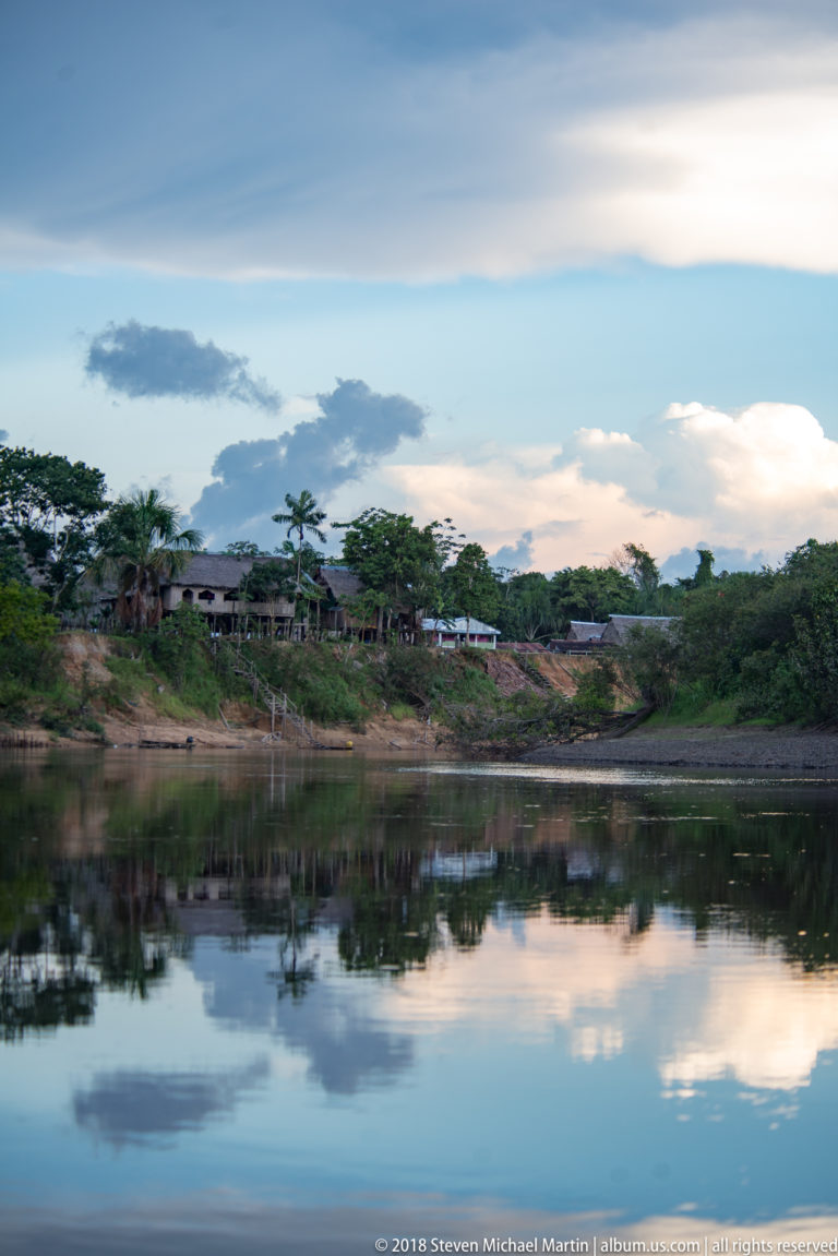Calm river reflecting blue-gray clouds, with a small riverside settlement and exposed bank across the water.