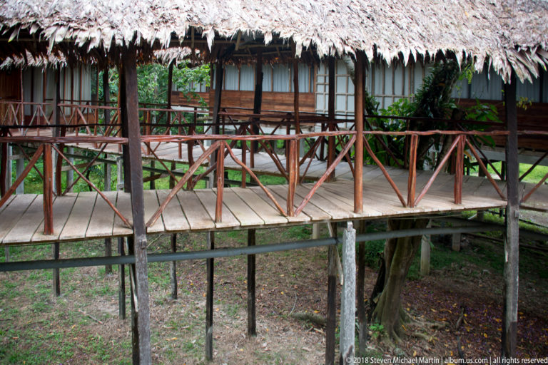 Raised wooden porch with rustic reddish railings and screened guest-room openings beneath deep palm-thatched eaves.