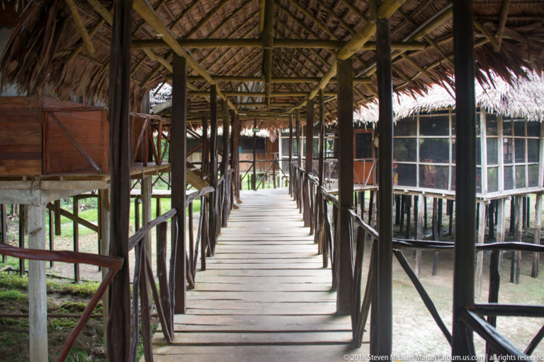 Covered wooden boardwalk running between stilted lodge buildings with screened walls and palm-thatched roofing.