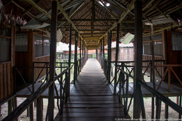 Straight covered wooden boardwalk between lodge rooms, with timber railings and a bright opening at the far end.