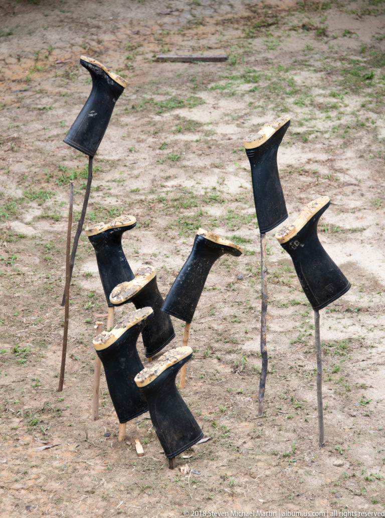Cluster of dark rustic wooden seats with narrow supports and curved saddle-like tops standing on sandy ground.