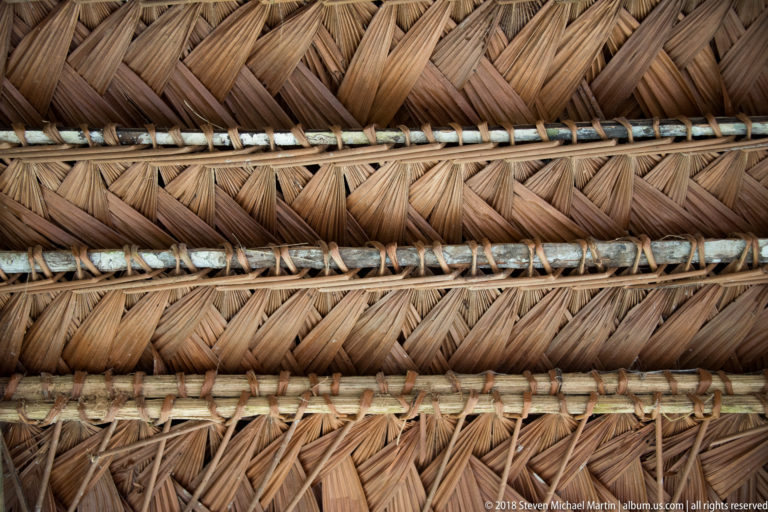 Close view of tightly woven palm-thatch roofing fastened in overlapping rows to horizontal wooden poles with lashings.