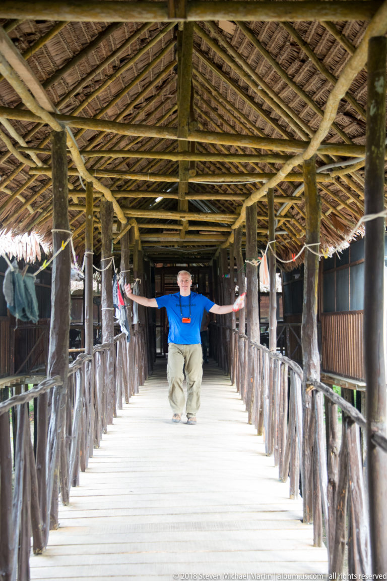 Visitor standing with arms outstretched in a long palm-thatched wooden corridor lined with rustic railings and lodge rooms.