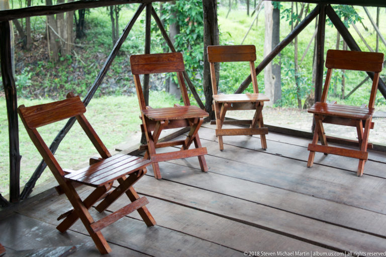 Four wooden folding chairs arranged on a plank-floored screened veranda with green forest visible beyond.