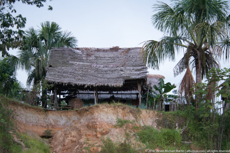 Open-sided palm-thatched shelter standing above an eroded sandy riverbank between tall palm trees.