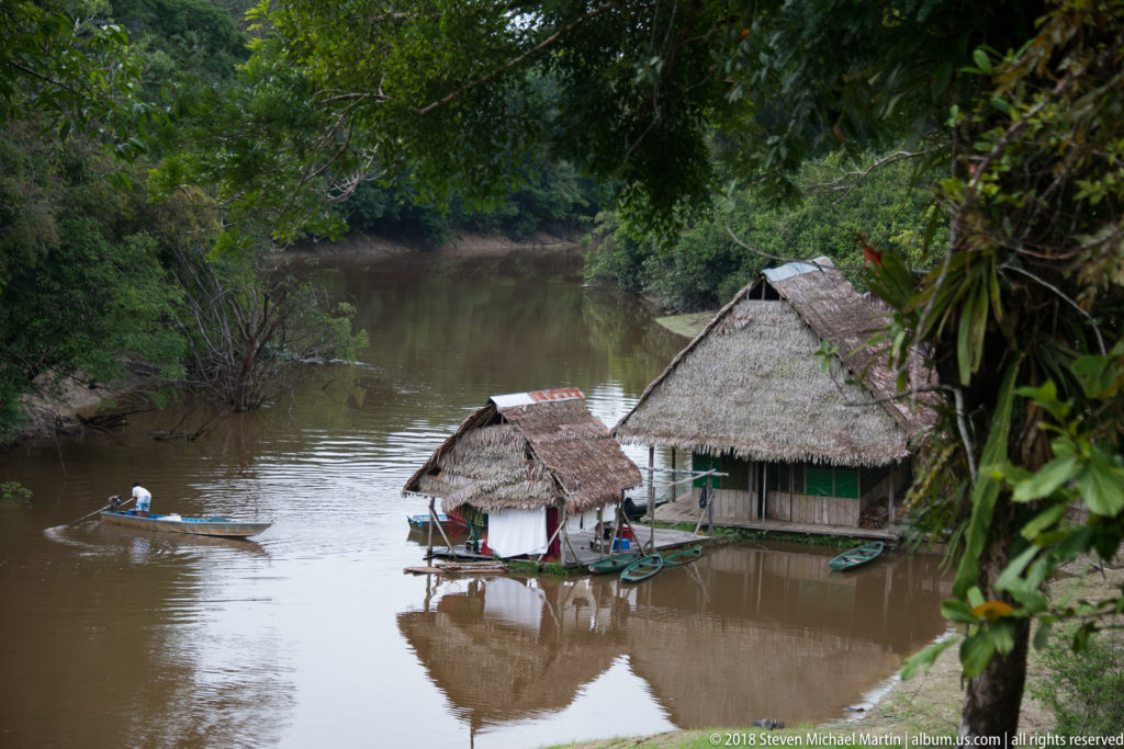 Palm-thatched wooden buildings on stilts beside a muddy Amazonian channel, with small boats and a motorized canoe nearby.