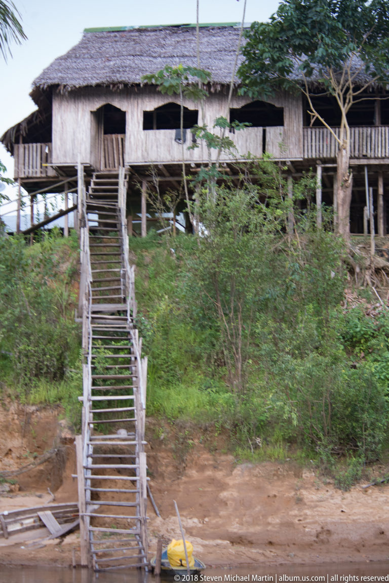 Long raised wooden house with veranda above a steep riverbank and narrow timber stairs descending toward the water.