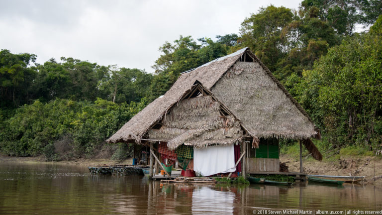 Steep thatched-roof dwelling at waterline beside a forested riverbank near Tuhauyo Lodge.