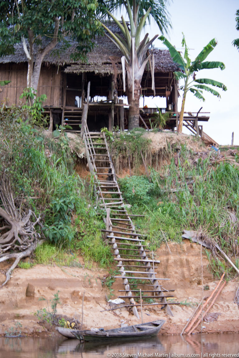 Palm-thatched stilt house above an eroded riverbank with a narrow wooden stairway descending to a canoe at the water.