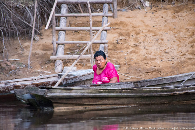 Woman in a bright pink shirt seated in a weathered wooden canoe below a rough stairway on the sandy bank.