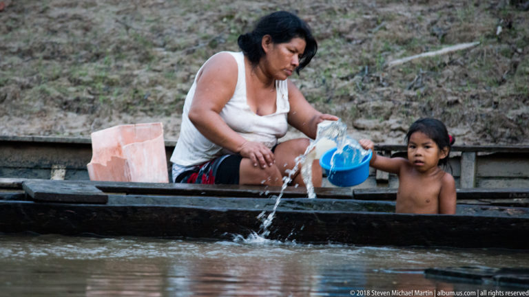 Adult and child in a wooden canoe pouring water from blue plastic containers over the side at the riverbank.