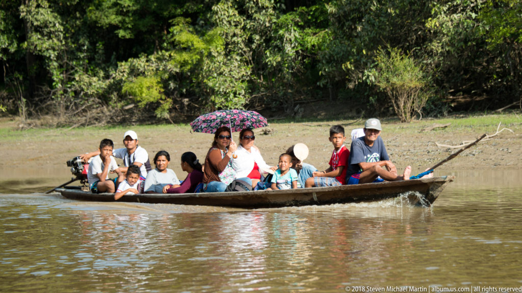 Adults and children crowded into a long wooden boat, with a patterned umbrella raised for shade.