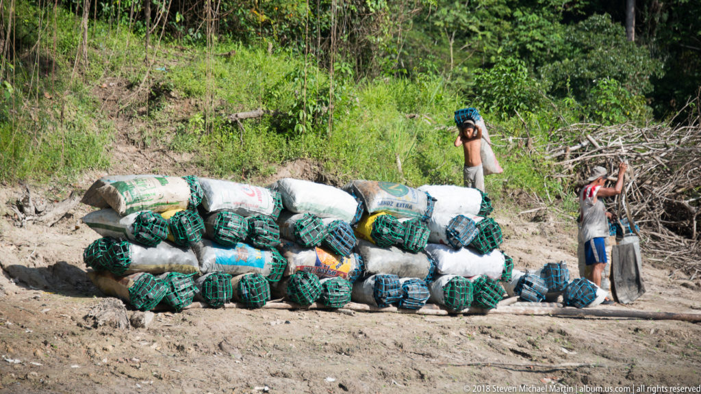 Large pile of sacks and green-tied bundles on a muddy riverbank with two people standing nearby.