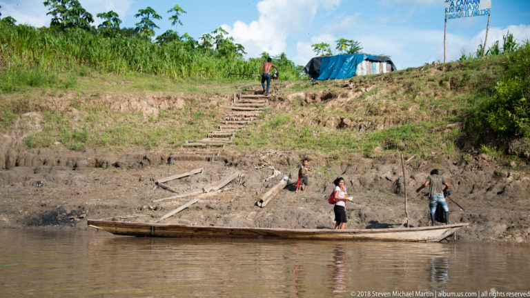 People with a long wooden canoe at the base of rough steps climbing a steep muddy riverbank.