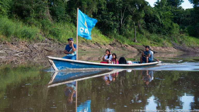 Family in a long narrow boat with a blue flag, reflected in calm brown river water.