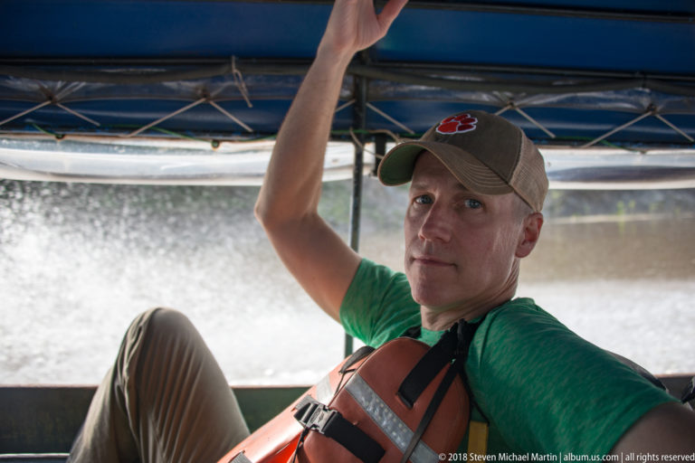 Seated passenger in an orange life jacket holding the canopy frame inside a moving riverboat.