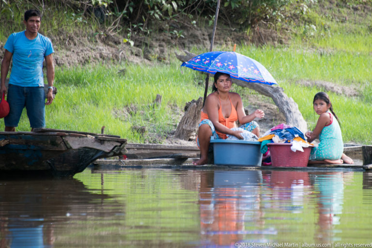 Woman and young girl seated beside laundry tubs under a blue umbrella at the riverbank.