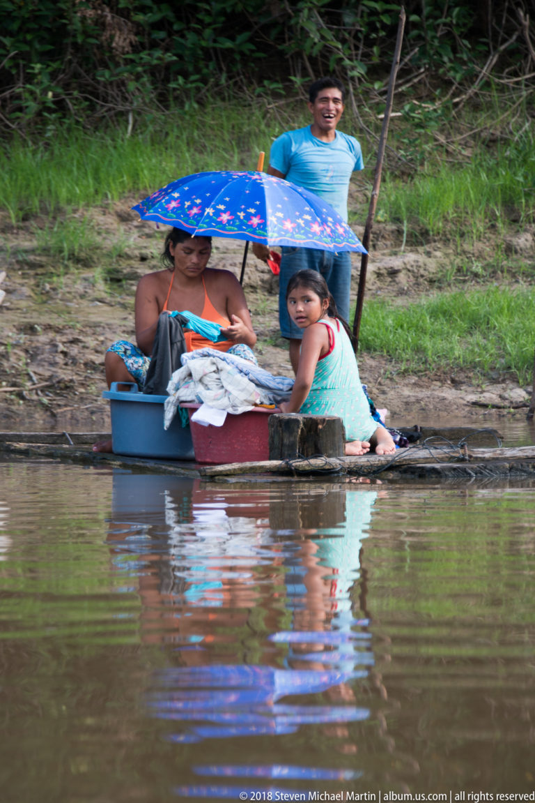 Woman and child beside tubs of laundry under a blue umbrella at the river edge, with a man behind.