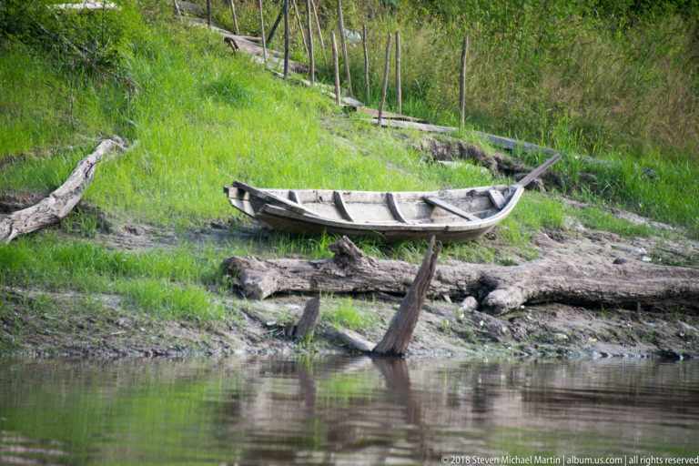 Long weathered wooden canoe resting on a grassy, muddy riverbank among driftwood and fence posts.