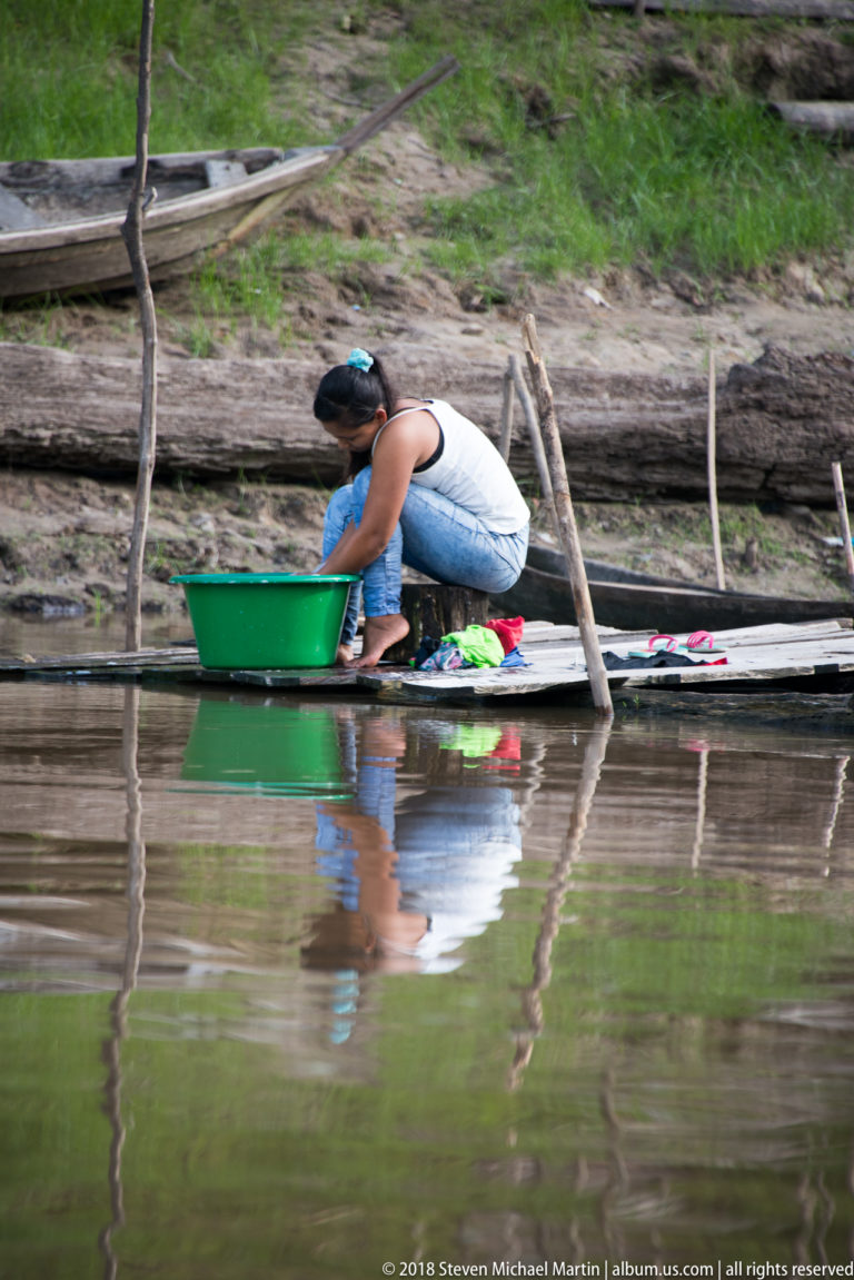 Woman crouching in a narrow canoe beside a green basin and clothing at the river edge.