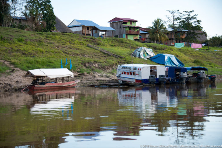 Several covered and open boats moored below houses on a grassy Tuhauyo River bank.
