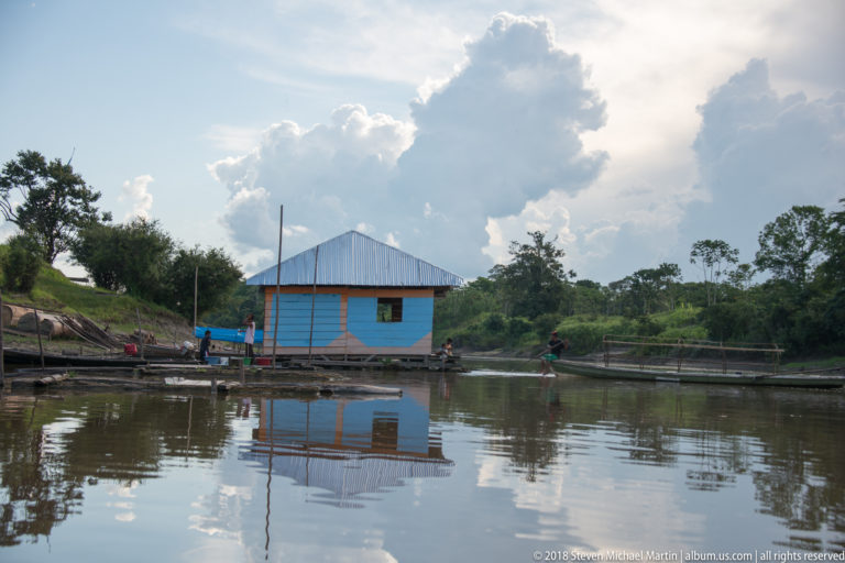 Small blue floating house reflected in calm river water beneath large white cumulus clouds.