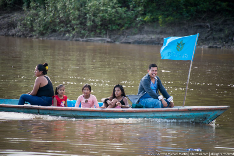 Adults and children seated in a long narrow boat with a blue flag on the Tuhauyo River.