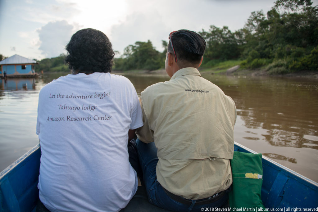Two passengers seen from behind in a blue boat traveling along the brown Tuhauyo River.
