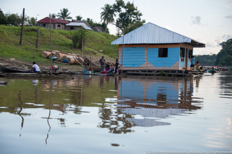 Blue floating riverside house surrounded by residents and narrow canoes, reflected in calm water.