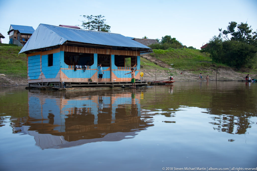 Blue wooden floating house with metal roof, porch, people, and a clear reflection in brown river water.