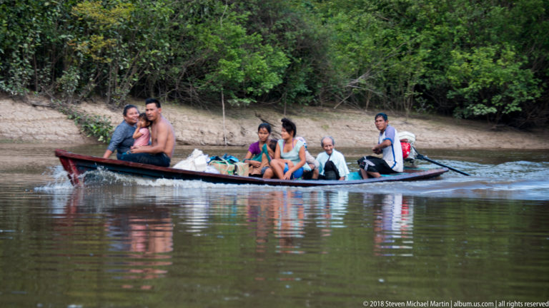 Six passengers and supplies ride in a narrow wooden motorboat along a calm forest-lined river.