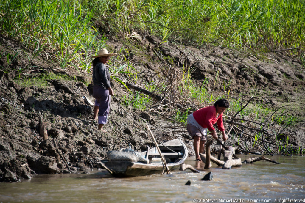 Two women beside a small canoe on a muddy riverbank, one cutting a fallen branch with a machete.