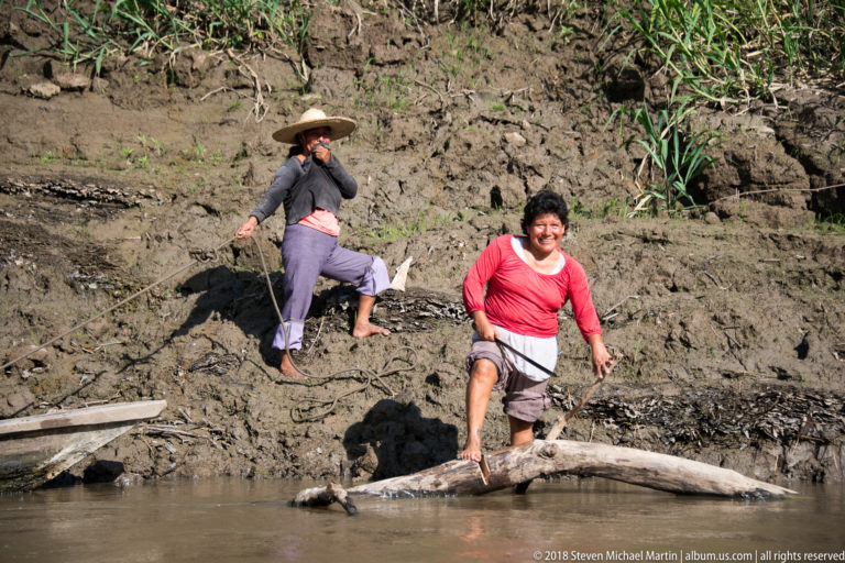 Two women descend a steep muddy riverbank, one stepping onto a fallen trunk at the brown river’s edge.