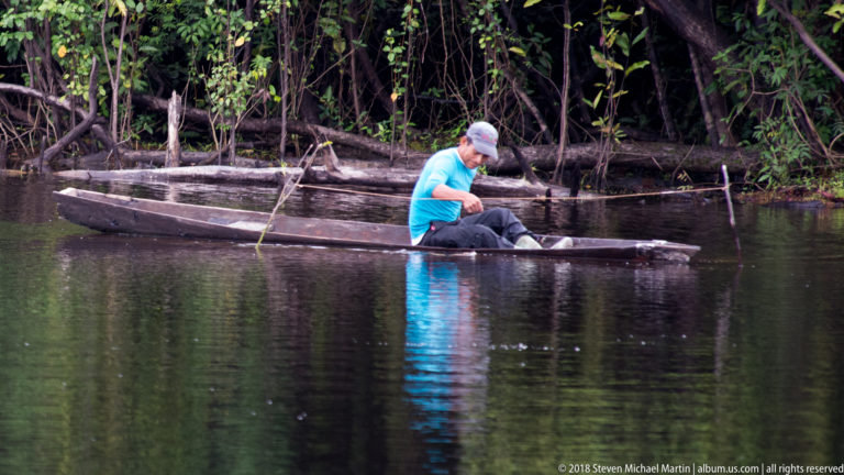 Person in turquoise shirt paddles a narrow wooden canoe beside another weathered boat at the forest edge.