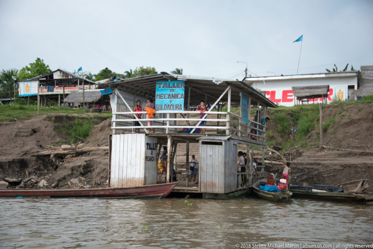 Two-level riverside workshop with people on the upper deck and narrow canoes tied along the muddy bank.