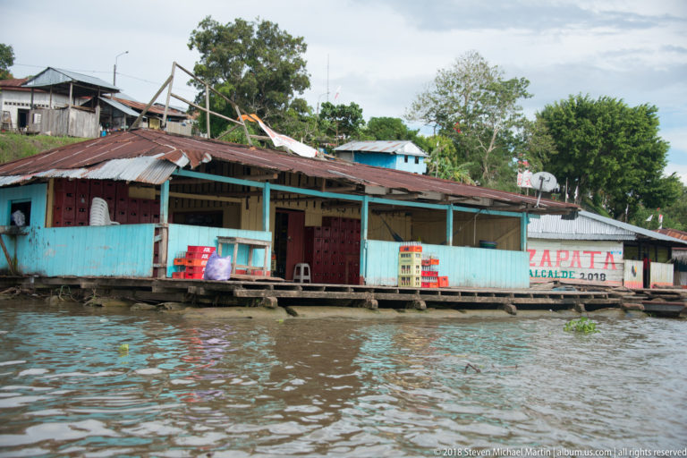 Blue wooden riverside shopfront with open veranda, corrugated roof, drink crates, and a painted sign beside the water.