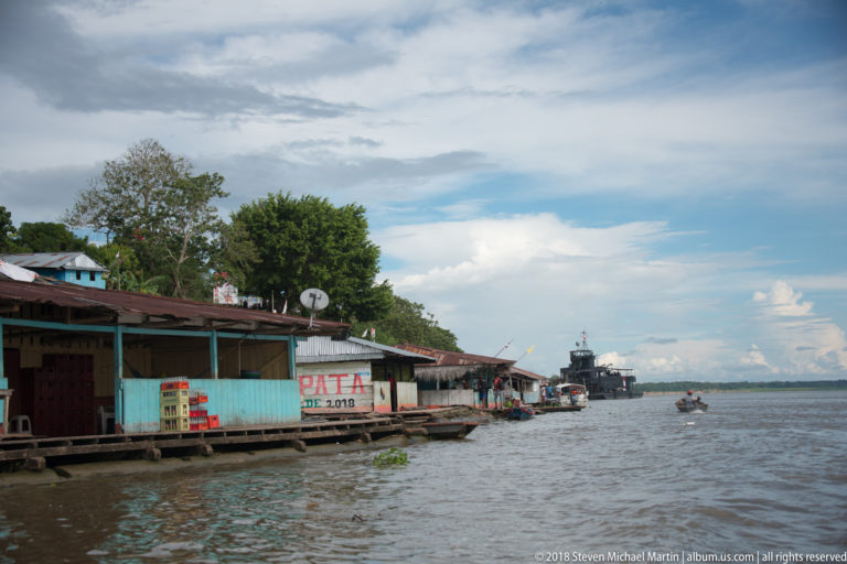 Row of low wooden riverside shops and houses with corrugated roofs, signs, and small boats along the channel.