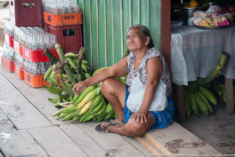 Older woman sits beside large green plantain bunches at a small shop with drink crates and packaged goods.