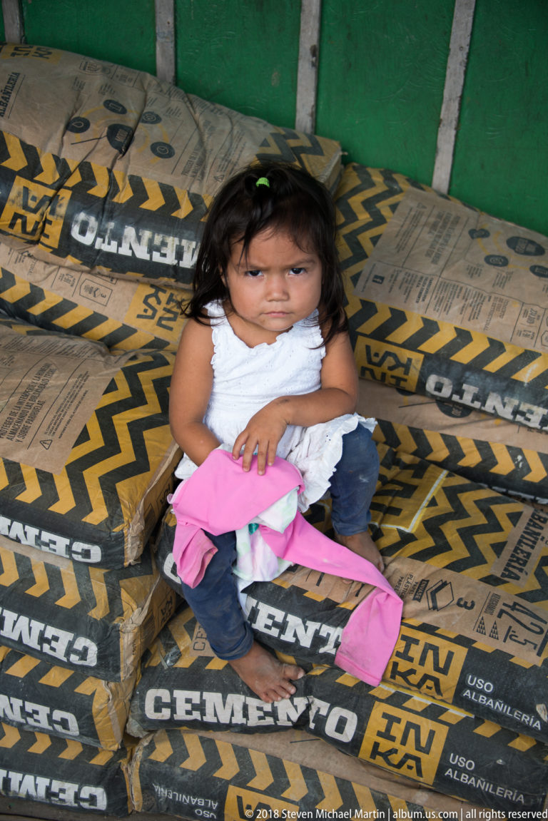 Young child in a white top sits on stacked black-and-yellow cement bags while holding a pink garment.