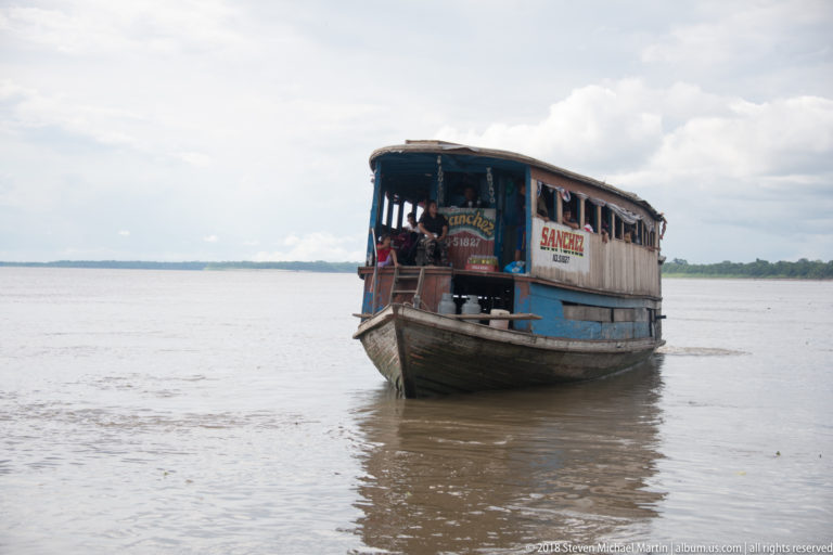 Weathered wooden regional riverboat approaches across a wide muddy channel under a cloudy sky.