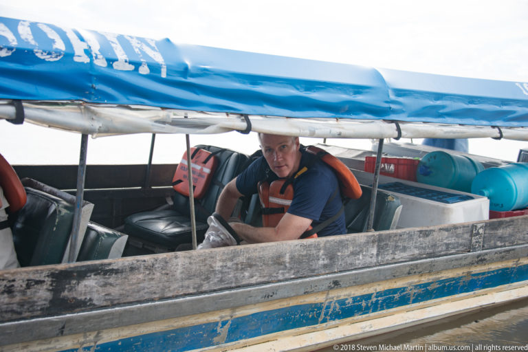 Close view of a man in a flotation vest inside a narrow motorboat beneath a rolled blue canopy.